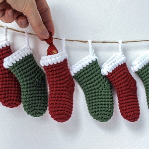 May include: A string of miniature crocheted stockings in red and green with white trim, hanging from a twine string. A hand is reaching into one of the stockings to retrieve a small chocolate bar.