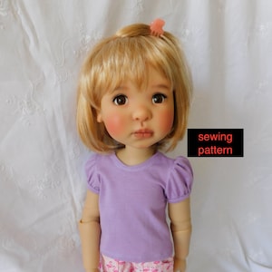 May include: A doll wearing a lavender t-shirt with puffed sleeves and pink flamingo print shorts. The doll has blonde hair and brown eyes. The text "sewing pattern" is visible in the image.