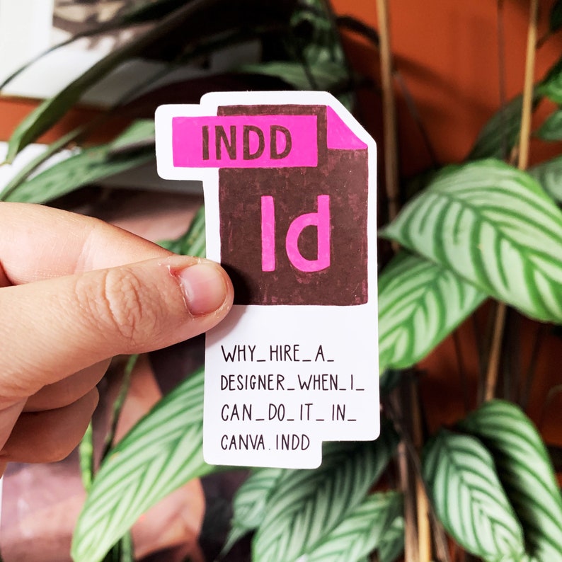 K&ouml;nnte beinhalten: Ein wei&szlig;er Aufkleber mit einem rosa und braunen InDesign-Datei-Symbol und dem Text "WHY_HIRE_A_DESIGNER_WHEN_I_CAN_DO_IT_IN_CANVA. INDD"