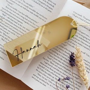Op de afbeelding: Een gouden bladwijzer met de naam "Hannah" geschreven in zwarte schrijfletters. De bladwijzer heeft de vorm van een hart en heeft een reflecterend oppervlak.