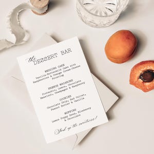 May include: A dessert bar menu on a white card with a list of treats, including wedding cake, French macarons, cookies, and muffins. The menu is next to an apricot, a cut apricot, a glass, and a spool of ribbon.