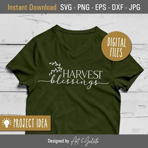 May include: A green t-shirt with a white design that says "HARVEST blessings" with a small floral design above the text. The text is in a script font. The t-shirt is on a white wood background.