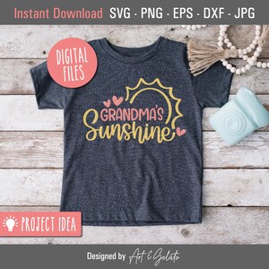 May include: A gray t-shirt with the text "Grandma's Sunshine" in yellow and pink lettering. The text is surrounded by a yellow sun and pink hearts. The t-shirt is on a wooden surface.