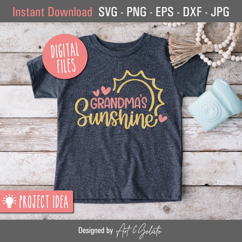 May include: A gray t-shirt with the text "Grandma's Sunshine" in yellow and pink lettering. The text is surrounded by a yellow sun and pink hearts. The t-shirt is on a wooden surface.