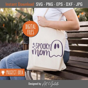 May include: A white tote bag with a purple ghost and the text "SPOOKY mom" on it. The bag is being held by a person wearing blue jeans and a white shirt.