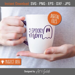 May include: White ceramic mug with a purple design that says "SPOOKY mom" with a ghost illustration. The mug is being held by a person's hands.