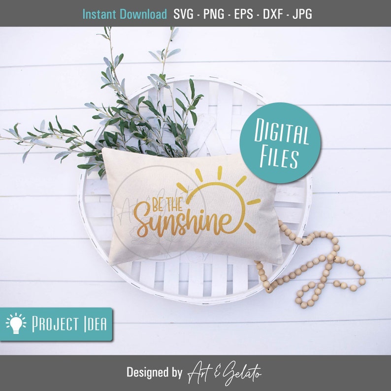 May include: A white pillow with the text "Be the Sunshine" in gold lettering and a yellow sun graphic. The pillow is sitting on a white wicker basket with a sprig of greenery.