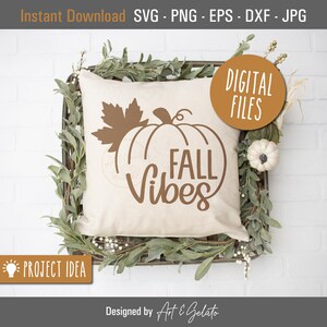 May include: A white pillow with a brown pumpkin and leaf design and the text "FALL Vibes". The pillow is sitting in a woven basket with greenery.