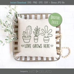 May include: A white pillow with a green line drawing of three potted plants and the text "LOVE GROWS HERE" in green. The pillow is sitting in a wooden tray with a white background.