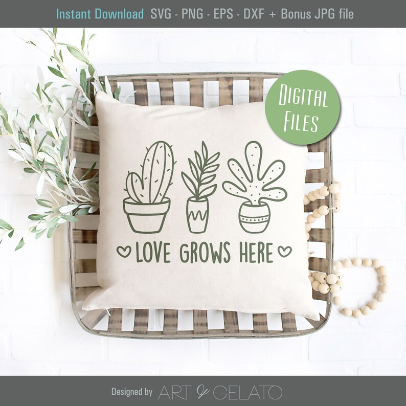 May include: A white pillow with a green line drawing of three potted plants and the text "LOVE GROWS HERE" in green. The pillow is sitting in a wooden tray with a white background.