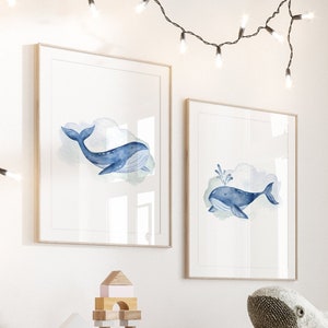 May include: Two framed watercolor prints of blue whales with light blue clouds. The prints are displayed on a white wall, with a string of lights hanging above. A white shelf below holds wooden blocks and a whale plush toy.