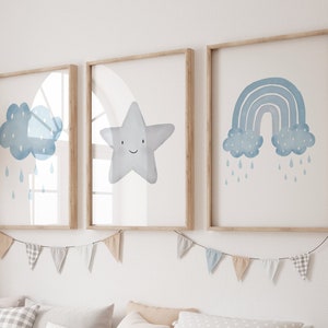 Puede incluir: Tres impresiones de acuarela enmarcadas con un esquema de color azul. Las impresiones presentan una nube con gotas de lluvia, una estrella sonriente y un arcoíris con nubes y gotas de lluvia. Las impresiones están colgadas en una pared blanca con una guirnalda azul claro y blanca.