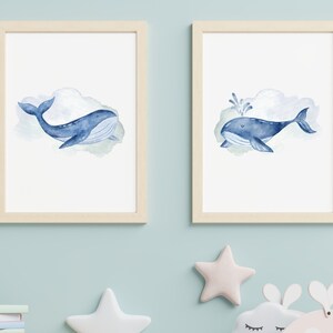 May include: Two framed watercolor prints featuring blue whales. One shows a whale swimming, the other with a water spout. Both whales are set against a soft, watercolor cloud background. The frames are light wood.