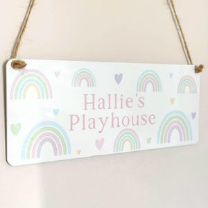 May include: A rectangular white sign with the text "Hallie's Playhouse" in pink, surrounded by pastel rainbows and hearts. The sign is suspended by a natural twine rope, perfect for a child's room or play area.