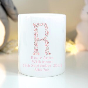 May include: White ceramic mug featuring a floral letter R design. The mug is personalised with the name Rosie Anne Wilkinson, the date 17th September 2024, and the weight 8lbs 7oz.