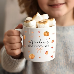 May include: A white ceramic mug filled with toasted marshmallows, featuring a printed design of orange pumpkins, autumn leaves, small hearts, and bows. The text on the mug reads "Amelia's HOT CHOCOLATE" in a combination of script and block lettering.