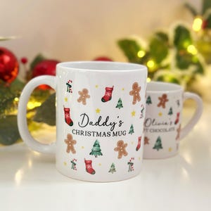 K&ouml;nnte beinhalten: Zwei wei&szlig;e Keramik-Weihnachtstassen. Auf der gr&ouml;&szlig;eren Tasse steht "Daddy's Christmas Mug" und sie ist mit Lebkuchenm&auml;nnern, Weihnachtsb&auml;umen und Str&uuml;mpfen verziert. Auf der kleineren Tasse steht "Olivia's Hot Chocolate".