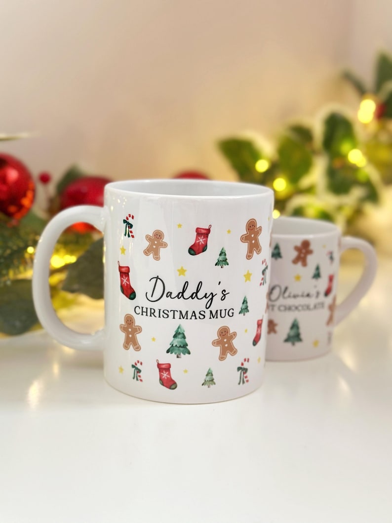 K&ouml;nnte beinhalten: Zwei wei&szlig;e Keramik-Weihnachtstassen. Auf der gr&ouml;&szlig;eren Tasse steht "Daddy's Christmas Mug" und sie ist mit Lebkuchenm&auml;nnern, Weihnachtsb&auml;umen und Str&uuml;mpfen verziert. Auf der kleineren Tasse steht "Olivia's Hot Chocolate".