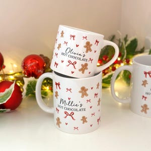 Könnte beinhalten: Zwei weiße Keramikbecher, dekoriert mit roten Schleifen, Lebkuchenmännern und Herzen, sind aufeinander gestapelt. Die Becher tragen die Aufschrift "Millie's HOT CHOCOLATE" und "Olivia's HOT CHOCOLATE". Festliches, weihnachtliches Design.