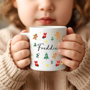 K&ouml;nnte beinhalten: Wei&szlig;e Keramiktasse mit weihnachtlichen Illustrationen, darunter Lebkuchenm&auml;nner, Weihnachtsb&auml;ume und Str&uuml;mpfe. Der Name "Freddie" ist in Schreibschrift geschrieben. Die Tasse wird von einer Person gehalten, die einen beigen Pullover tr&auml;gt.