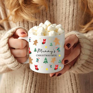 K&ouml;nnte beinhalten: Wei&szlig;e Keramik-Weihnachtstasse gef&uuml;llt mit Marshmallows, mit Lebkuchenm&auml;nnern, Weihnachtsb&auml;umen und Str&uuml;mpfen. Die Tasse tr&auml;gt den Text "Mummy's CHRISTMAS MUG" und gelbe Sterne. Ein festliches Weihnachtsgeschenk.