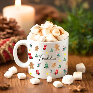 K&ouml;nnte beinhalten: Eine wei&szlig;e Keramiktasse gef&uuml;llt mit ger&ouml;steten Marshmallows, verziert mit weihnachtlichen Illustrationen. Die Tasse zeigt Lebkuchenm&auml;nner, Weihnachtsb&auml;ume, Zuckerstangen und Str&uuml;mpfe. Der Name "Freddie" ist auf der Tasse geschrieben.