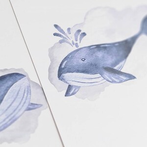May include: Watercolor illustration of a blue whale with a water spout, set against a soft gray cloud. The whale is depicted in shades of blue, with a detailed fin and tail. The artwork is on a white background.