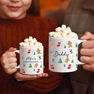 K&ouml;nnte beinhalten: Zwei wei&szlig;e Weihnachtstassen gef&uuml;llt mit Marshmallows. Auf einer Tasse steht "Alfie's CHRISTMAS MUG" und auf der anderen "Daddy". Beide Tassen haben Designs mit Weihnachtsb&auml;umen, Lebkuchenm&auml;nnern und Str&uuml;mpfen.