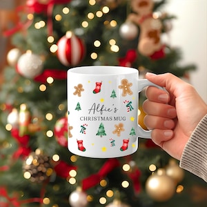K&ouml;nnte beinhalten: Wei&szlig;er Keramikbecher mit Henkel, verziert mit weihnachtlichen Illustrationen, darunter Lebkuchenm&auml;nner, Weihnachtsb&auml;ume und Str&uuml;mpfe. Der Becher tr&auml;gt den Text "Alfie's CHRISTMAS MUG" in schwarzer Schrift.