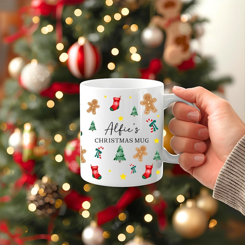 K&ouml;nnte beinhalten: Wei&szlig;er Keramikbecher mit Henkel, verziert mit weihnachtlichen Illustrationen, darunter Lebkuchenm&auml;nner, Weihnachtsb&auml;ume und Str&uuml;mpfe. Der Becher tr&auml;gt den Text "Alfie's CHRISTMAS MUG" in schwarzer Schrift.