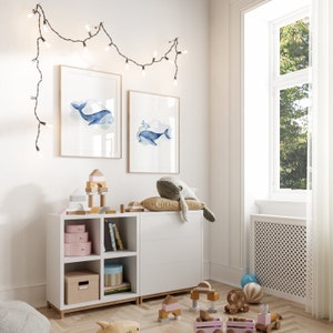 May include: A child's room with a white cabinet and shelves, adorned with framed whale art. A plush whale toy rests on the cabinet. Wooden blocks, toy train, and decorative boxes add to the playful decor. String lights hang above the artwork, creating a cozy atmosphere.