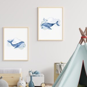 May include: Two framed watercolor prints featuring blue whales. One whale has a water spout. The whales are set against a soft, cloudy background. The frames are a light wood color. The prints are displayed in a child's room.