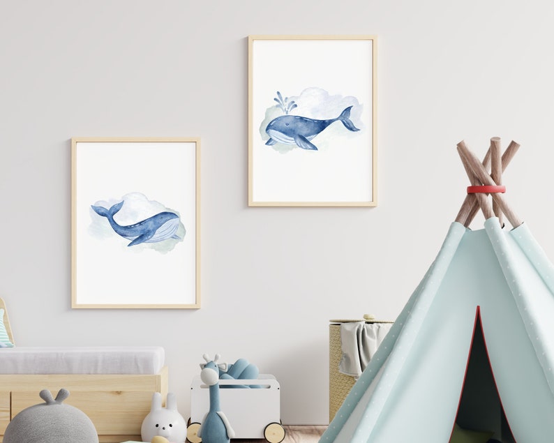 May include: Two framed watercolor prints featuring blue whales. One whale has a water spout. The whales are set against a soft, cloudy background. The frames are a light wood color. The prints are displayed in a child's room.