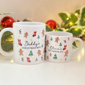 K&ouml;nnte beinhalten: Zwei wei&szlig;e Keramikbecher mit weihnachtlichen Designs. Der gr&ouml;&szlig;ere Becher tr&auml;gt die Aufschrift "Daddy's Christmas Mug" und der kleinere Becher "Olivia's Hot Chocolate". Beide Becher sind mit Lebkuchenm&auml;nnern, Weihnachtsb&auml;umen und Str&uuml;mpfen verziert.