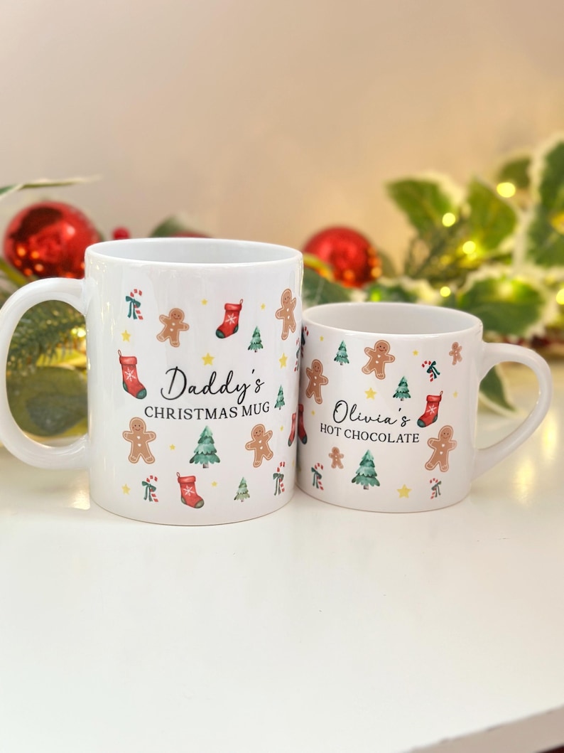 K&ouml;nnte beinhalten: Zwei wei&szlig;e Keramikbecher mit weihnachtlichen Designs. Der gr&ouml;&szlig;ere Becher tr&auml;gt die Aufschrift "Daddy's Christmas Mug" und der kleinere Becher "Olivia's Hot Chocolate". Beide Becher sind mit Lebkuchenm&auml;nnern, Weihnachtsb&auml;umen und Str&uuml;mpfen verziert.