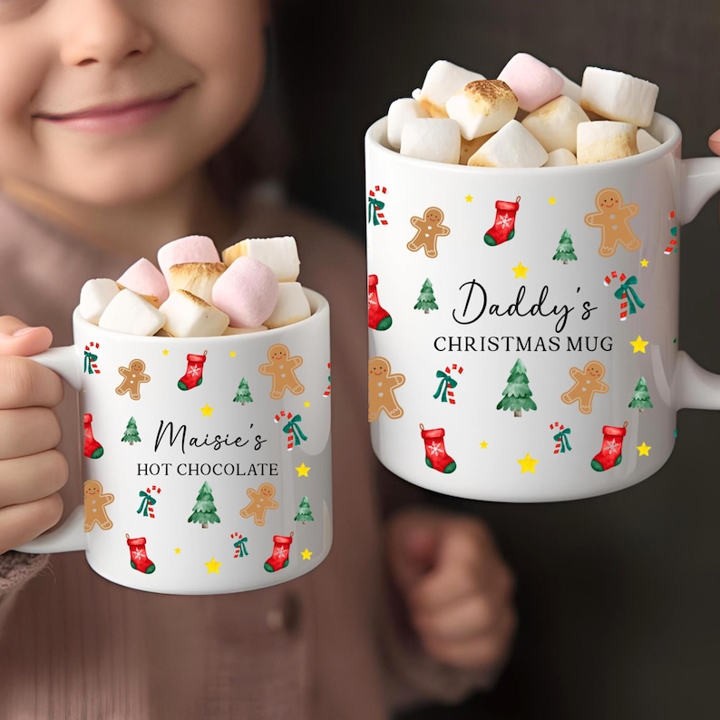 K&ouml;nnte beinhalten: Zwei wei&szlig;e Keramikbecher gef&uuml;llt mit Marshmallows. Auf einem Becher steht "Maisie's HOT CHOCOLATE" und auf dem anderen "Daddy's CHRISTMAS MUG". Beide Becher haben Lebkuchenm&auml;nner, Weihnachtsb&auml;ume, Str&uuml;mpfe und Sterne.