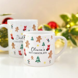 K&ouml;nnte beinhalten: Zwei wei&szlig;e Keramikbecher mit weihnachtlichen Designs. Auf einem Becher steht "Dad CHRISTMAS" und auf dem anderen "Olivia's HOT CHOCOLATE". Beide Becher zeigen Lebkuchenm&auml;nner, Weihnachtsb&auml;ume und rote Str&uuml;mpfe.