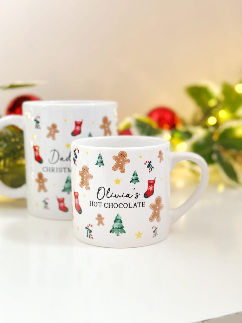K&ouml;nnte beinhalten: Zwei wei&szlig;e Keramikbecher mit weihnachtlichen Designs. Auf einem Becher steht "Dad CHRISTMAS" und auf dem anderen "Olivia's HOT CHOCOLATE". Beide Becher zeigen Lebkuchenm&auml;nner, Weihnachtsb&auml;ume und rote Str&uuml;mpfe.