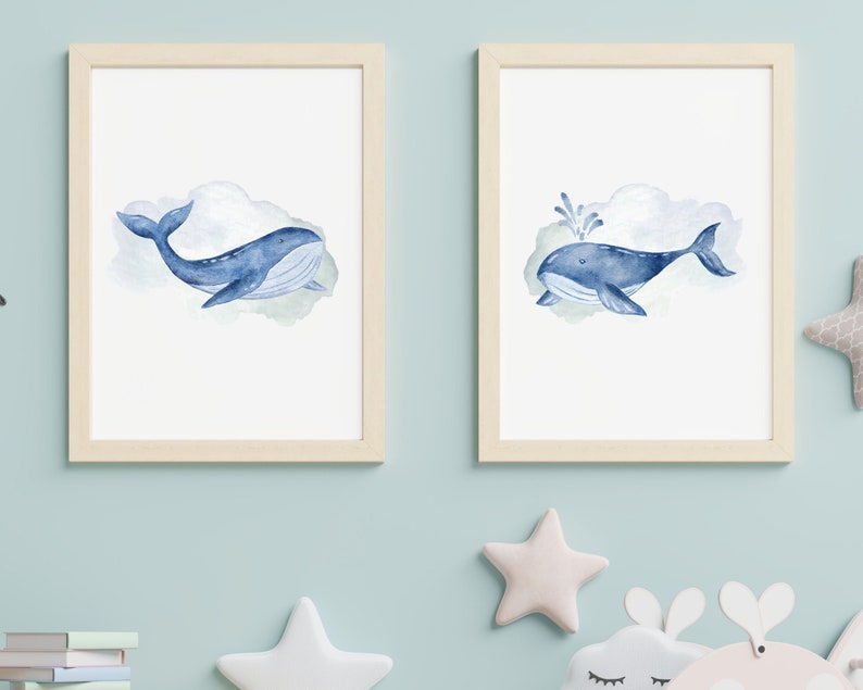 May include: Two framed watercolor prints featuring blue whales. One shows a whale swimming, the other with a water spout. Both whales are set against a soft, watercolor cloud background. The frames are light wood.