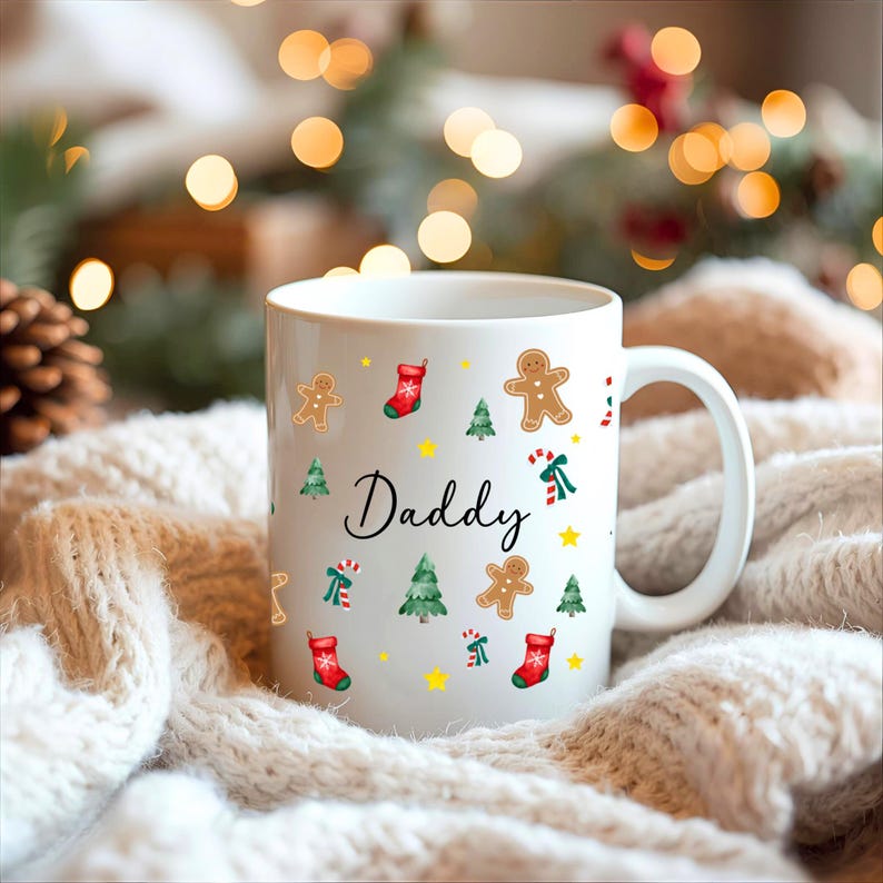 K&ouml;nnte beinhalten: Wei&szlig;e Keramik-Tasse mit dem Wort "Daddy" in Schreibschrift, verziert mit Lebkuchenm&auml;nnern, Weihnachtsb&auml;umen, Zuckerstangen, Sternen und roten Socken. Die Tasse hat einen wei&szlig;en Henkel.