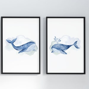 May include: Two framed watercolor prints featuring blue whales. One whale is swimming, the other is spouting water. Each whale is set against a soft, watercolor cloud background. The frames are black, and the art is on white paper.