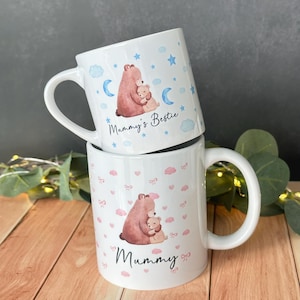 Könnte beinhalten: Zwei weiße Keramikbecher. Ein Becher zeigt einen braunen Bären, der einen kleineren beigen Bären umarmt, mit blauen Wolken, Sternen und Monden. Der Text "Mummy's Bestie" steht darunter. Der andere Becher hat ein ähnliches Design mit dem Text "Mummy" und rosa Schleifen.
