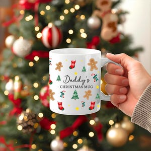 K&ouml;nnte beinhalten: Wei&szlig;e Keramik-Tasse mit Henkel, verziert mit weihnachtlichen Illustrationen, darunter Lebkuchenm&auml;nner, Weihnachtsb&auml;ume und Str&uuml;mpfe. Der Text "Daddy's CHRISTMAS MUG" ist aufgedruckt. Die Tasse wird vor einem Weihnachtsbaum gehalten.