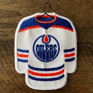 May include: White hockey jersey-shaped ornament with blue and orange detailing. The word "OILERS" is displayed in a circular emblem. Includes a black metal hanger for display.