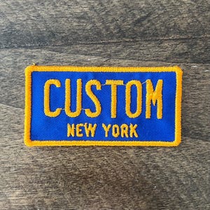 May include: Blue and yellow embroidered patch with the word "CUSTOM" and the text "NEW YORK" underneath.