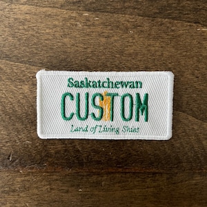 Peut inclure: Écusson brodé blanc avec du texte vert qui dit "Saskatchewan CUSTOM Land of Living Skies".