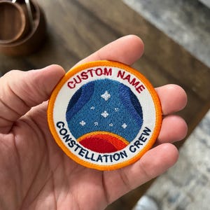 May include: A circular embroidered patch featuring a blue space-themed center with white stars and a red planet arc. The white border contains the red text CUSTOM NAME at the top and the blue text CONSTELLATION CREW at the bottom, with an orange edge.