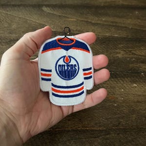 May include: A white hockey jersey patch with the Oilers logo in blue and orange. The jersey design includes blue and orange stripes on the sleeves and shoulders. A small black hanger is attached to the top of the patch.
