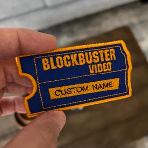 May include: A rectangular embroidered patch shaped like a movie ticket with a blue background and yellow border. The text BLOCKBUSTER VIDEO is embroidered in yellow at the top, and the words CUSTOM NAME are embroidered in yellow on a lower bar.
