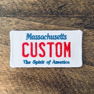 May include: White rectangular embroidered patch with the text "Massachusetts" in blue, "CUSTOM" in red, and "The Spirit of America" in blue. The patch is on a wooden surface.